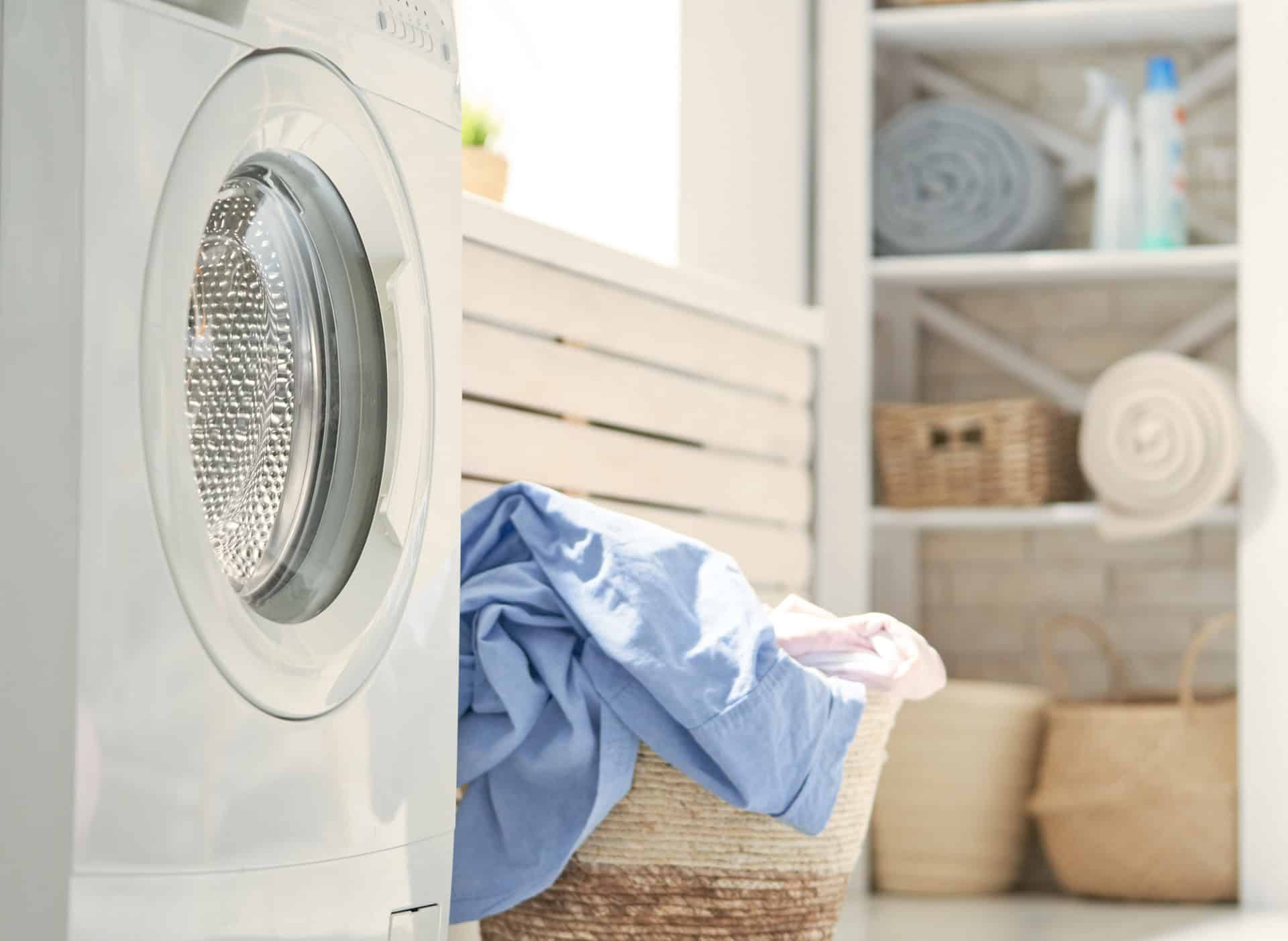 Front-loading washing machine next to a laundry basket filled with clothes, with shelves holding towels and baskets in the background.