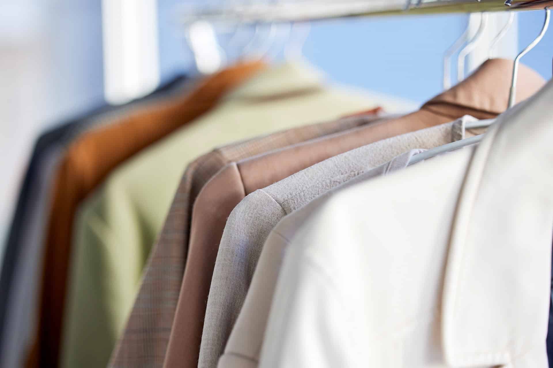 A row of assorted jackets and shirts in neutral colors hanging on white plastic hangers on a clothing rack.