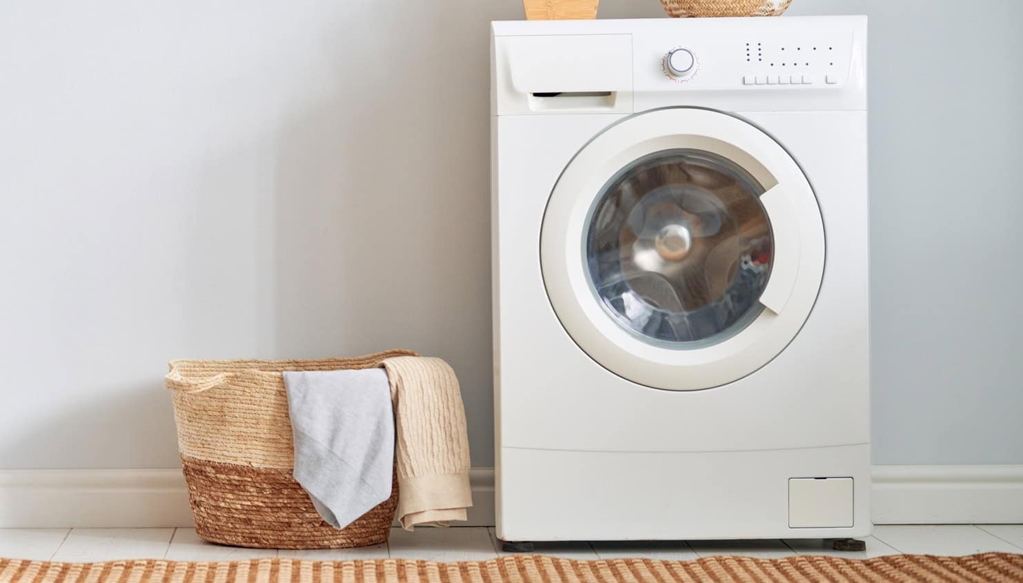 A front-loading washing machine next to a woven laundry basket with a towel draped over the side, set against a light-colored wall.