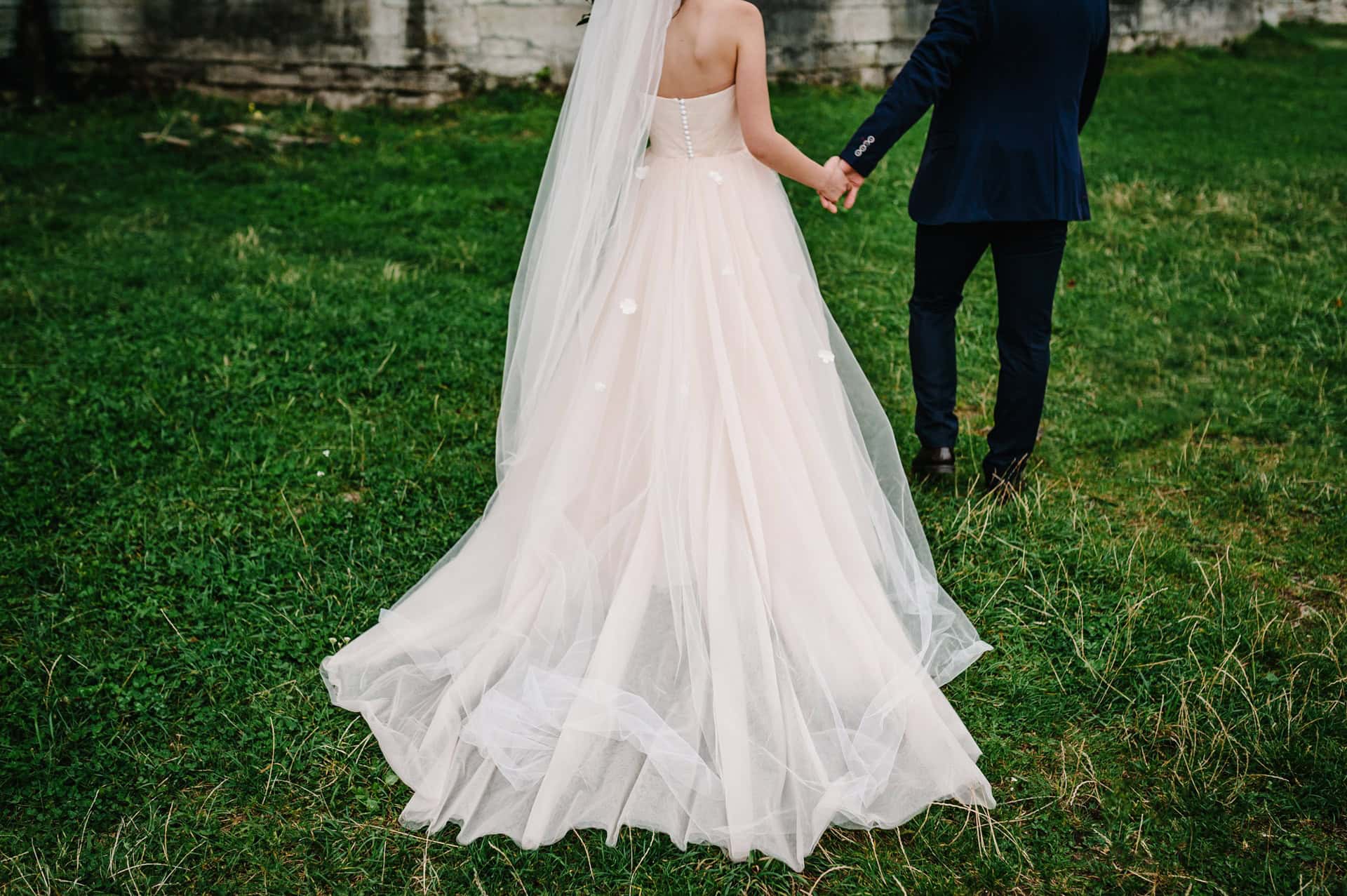 A bride in a white wedding dress and veil holds hands with a groom in a dark suit while standing on grass.