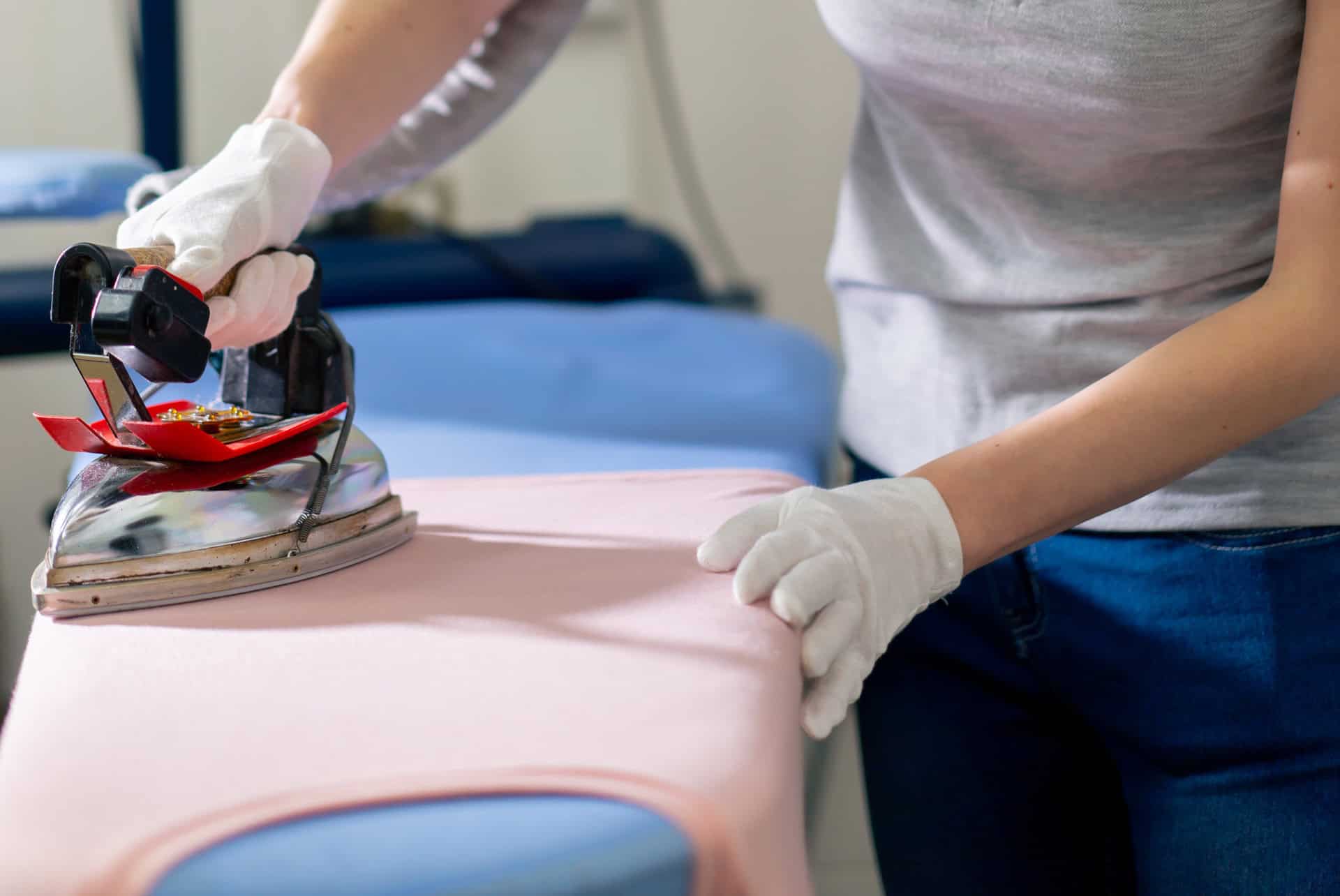 A person wearing white gloves is ironing a pink garment on an ironing board, holding the fabric taut with one hand and pressing with an iron in the other hand.