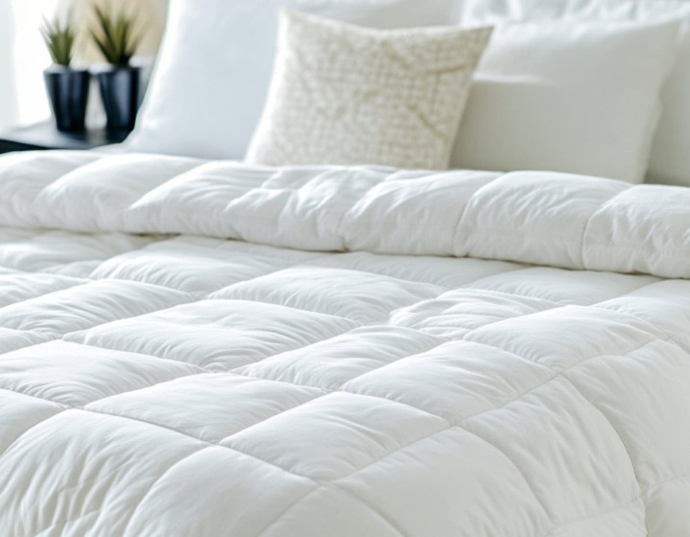 Close-up of a neatly made bed with a white quilted comforter and several white pillows, with two small potted plants in the background.
