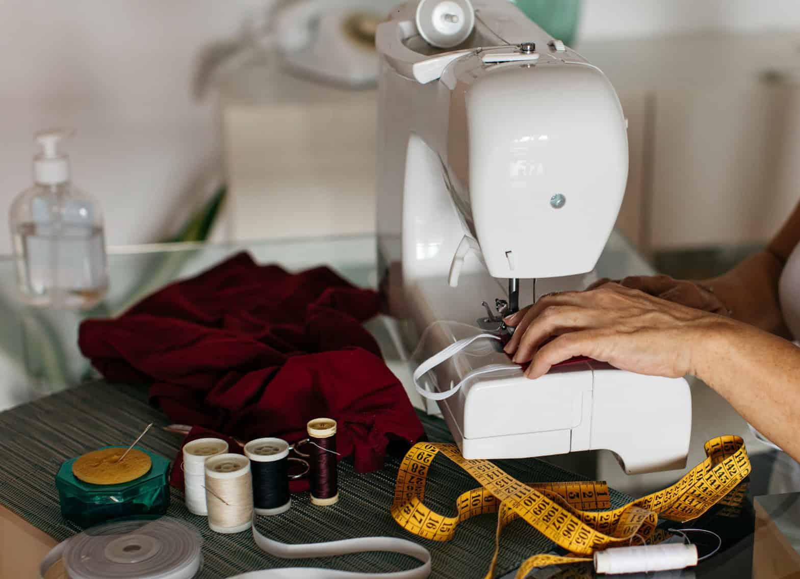 Person using a sewing machine with various sewing supplies, including thread spools, tape measure, fabric, and pins, arranged on a table.