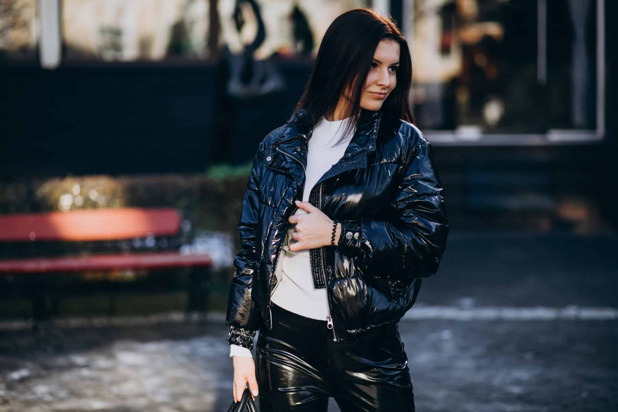 A woman wearing a shiny black jacket and pants stands outdoors on a paved area, looking to the side with one hand holding her jacket.