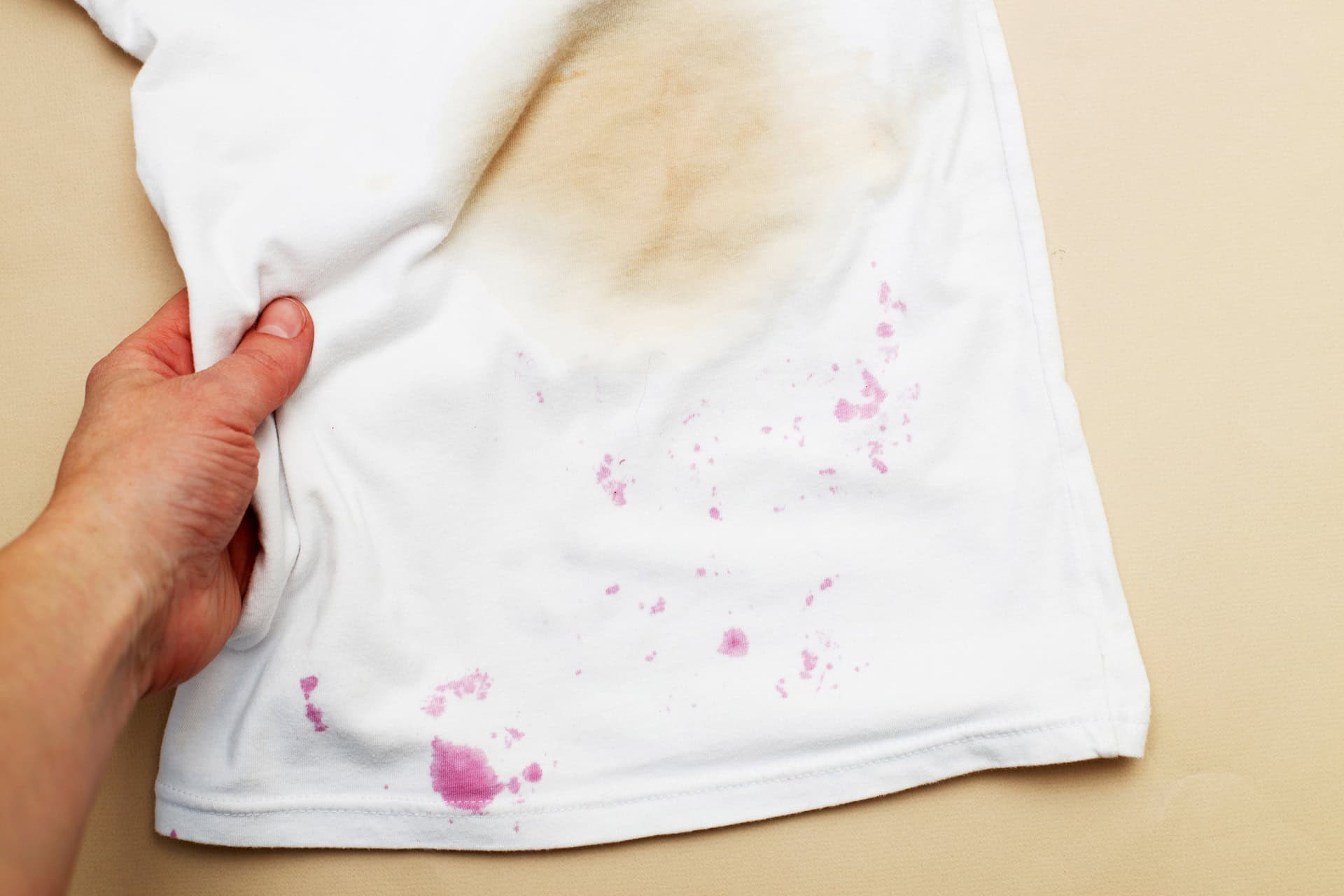 A hand holds a white shirt with a large brown stain and several smaller purple stains on the fabric.