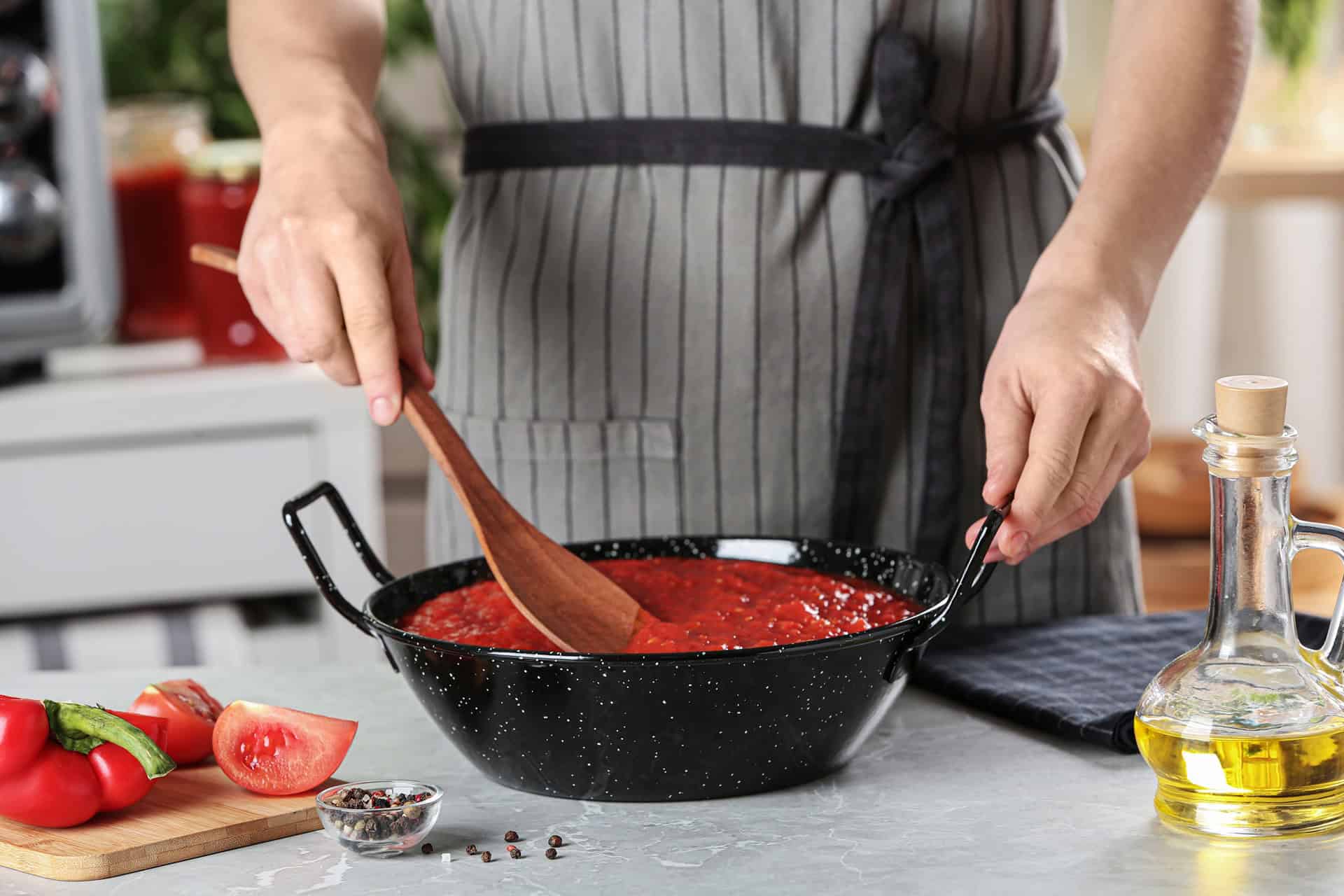 Person stirring tomato sauce in a black speckled pan with a wooden spoon; fresh tomatoes, spices, and olive oil on the counter nearby.