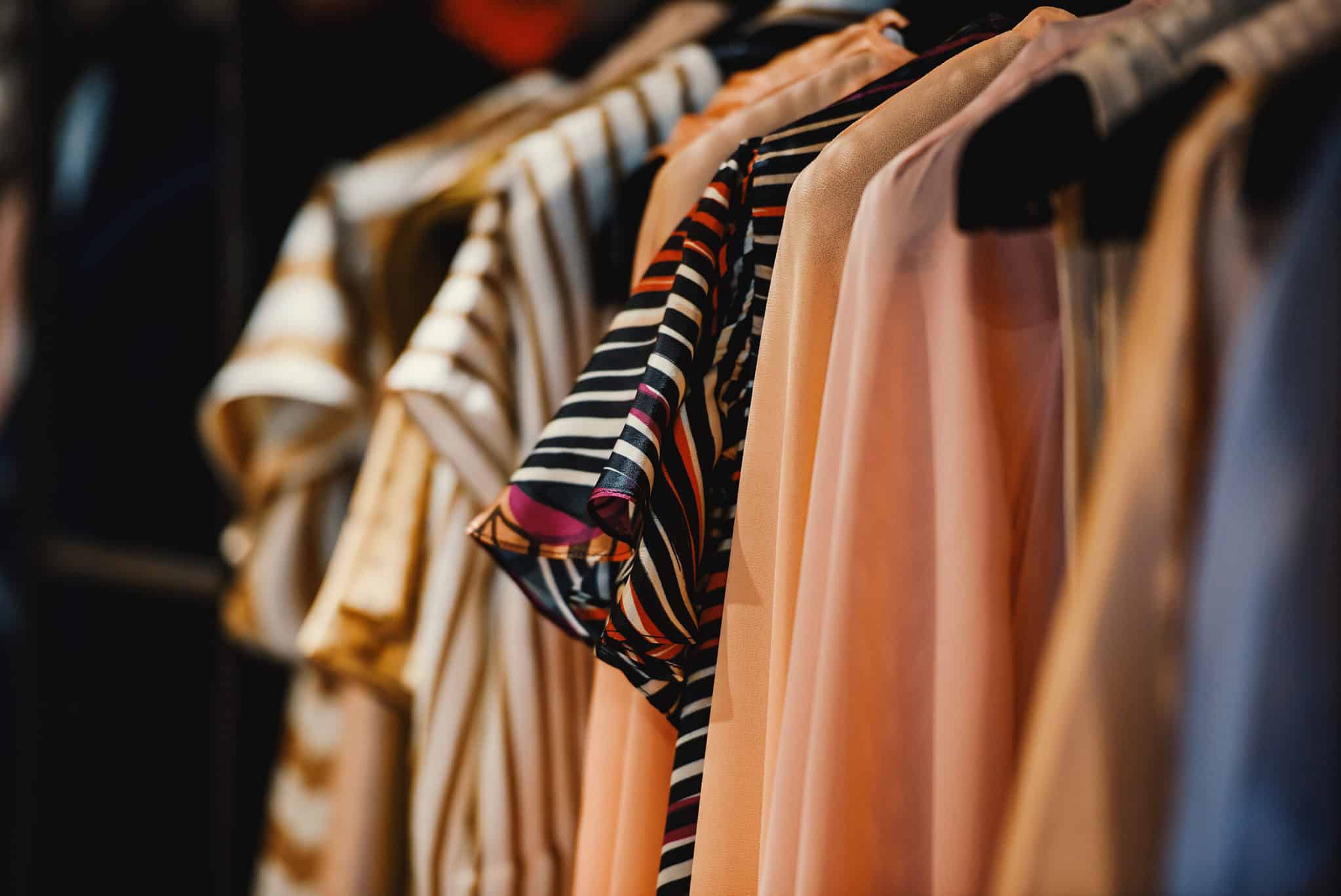 A row of various shirts and blouses, including striped and solid-colored patterns, hanging on black hangers in a closet or store.