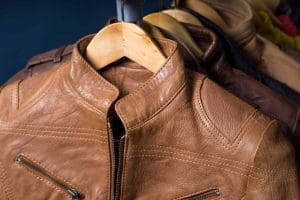 A close-up of a light brown leather jacket on a wooden hanger, with other jackets hanging in the background.