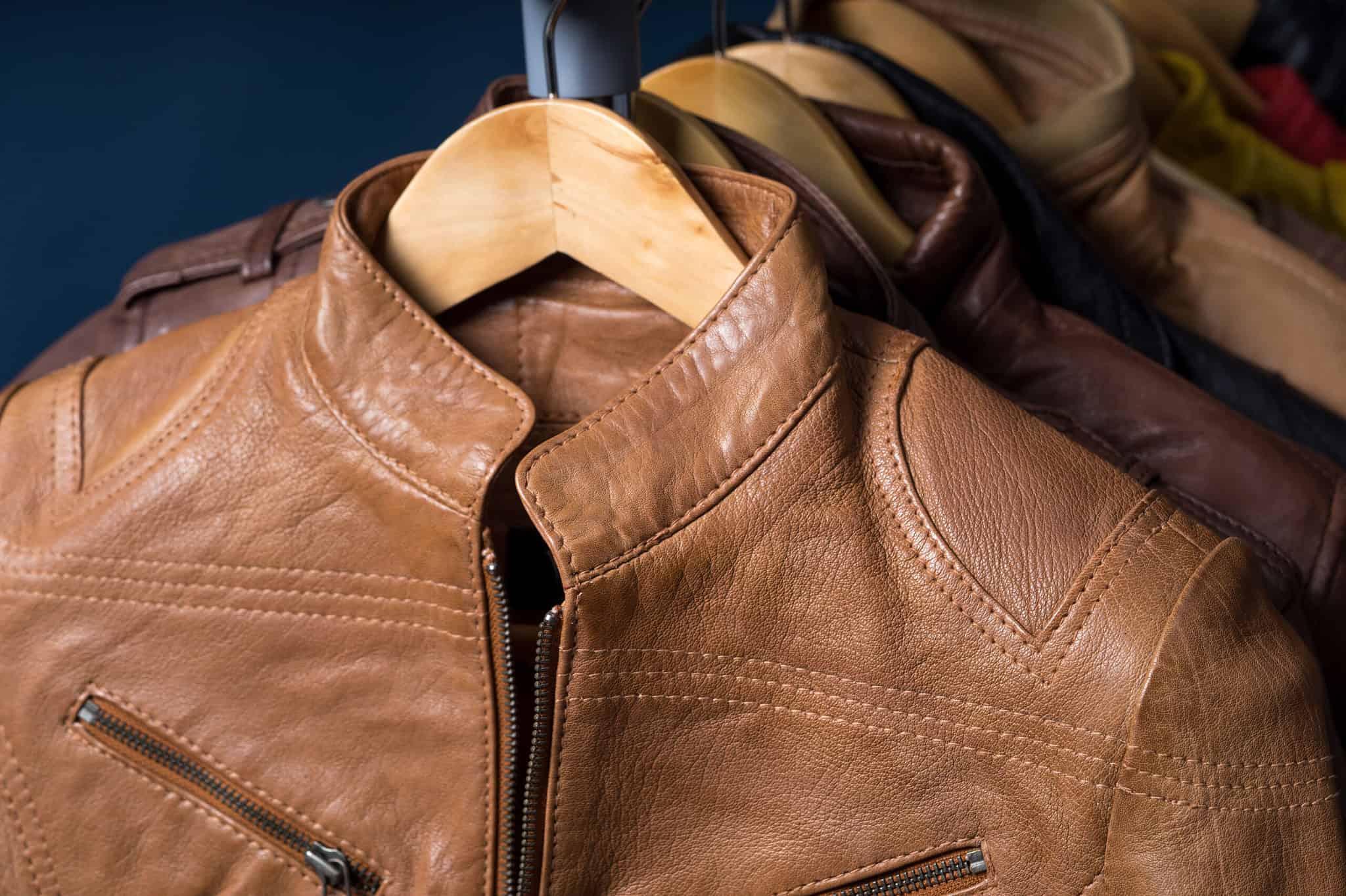 A close-up of a light brown leather jacket on a wooden hanger, with other jackets hanging in the background.