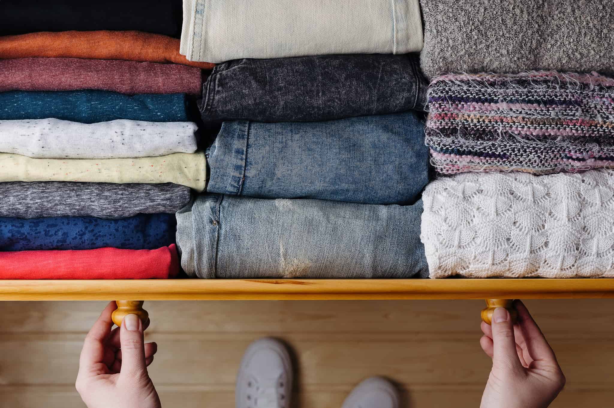 A person opens a wooden drawer filled with neatly folded clothes arranged by color and texture.