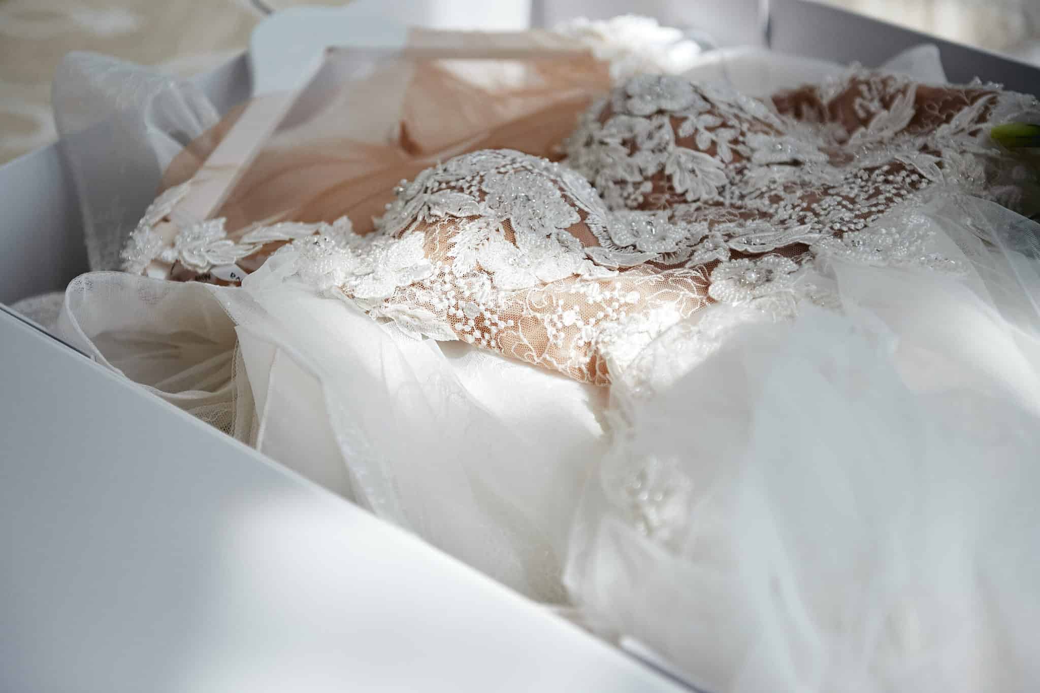 A white lace wedding dress with intricate beading and embroidery is folded inside a white box.