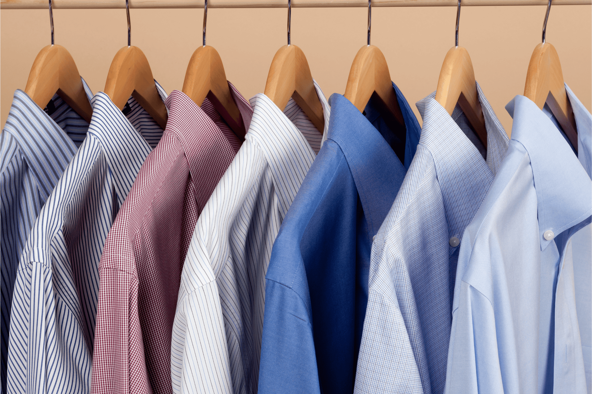 Seven collared dress shirts in various colors and patterns, including stripes and solid blue, hanging neatly on wooden hangers from a metal rod.