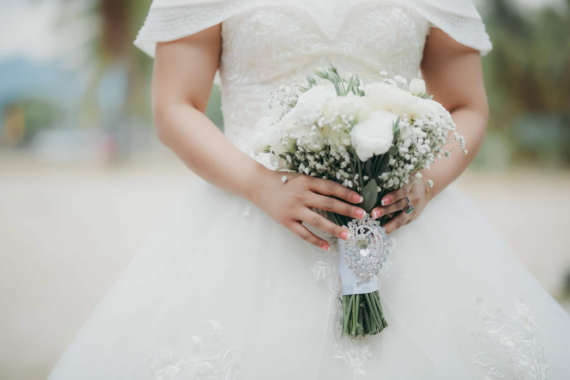 A bride in a white wedding dress holds a bouquet of white flowers and greenery, with lace and bead details on the dress and bouquet.