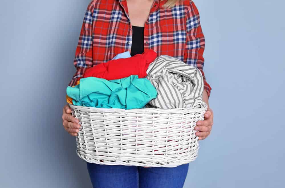 A person in a red plaid shirt holds a white woven basket filled with colorful clothes against a blue background.