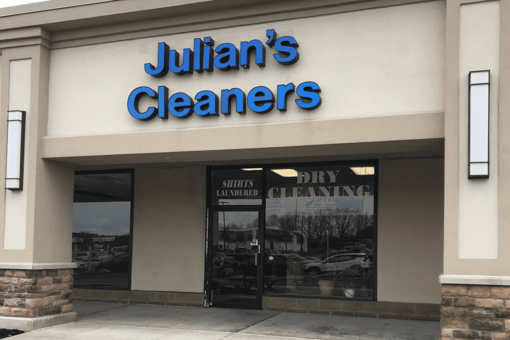 Storefront of "Julian’s Cleaners" with blue signage above glass doors and windows, showing reflections and interior signs for shirts laundered and dry cleaning.