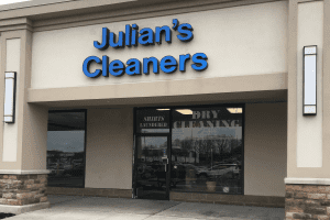 Storefront of "Julian&rsquo;s Cleaners" with blue signage above glass doors and windows, showing reflections and interior signs for shirts laundered and dry cleaning.