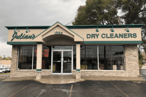 A one-story building with a sign reading "Julian's Dry Cleaners 1964" and a "Same Day Service" neon sign in the window. The exterior features stone accents and large glass windows.