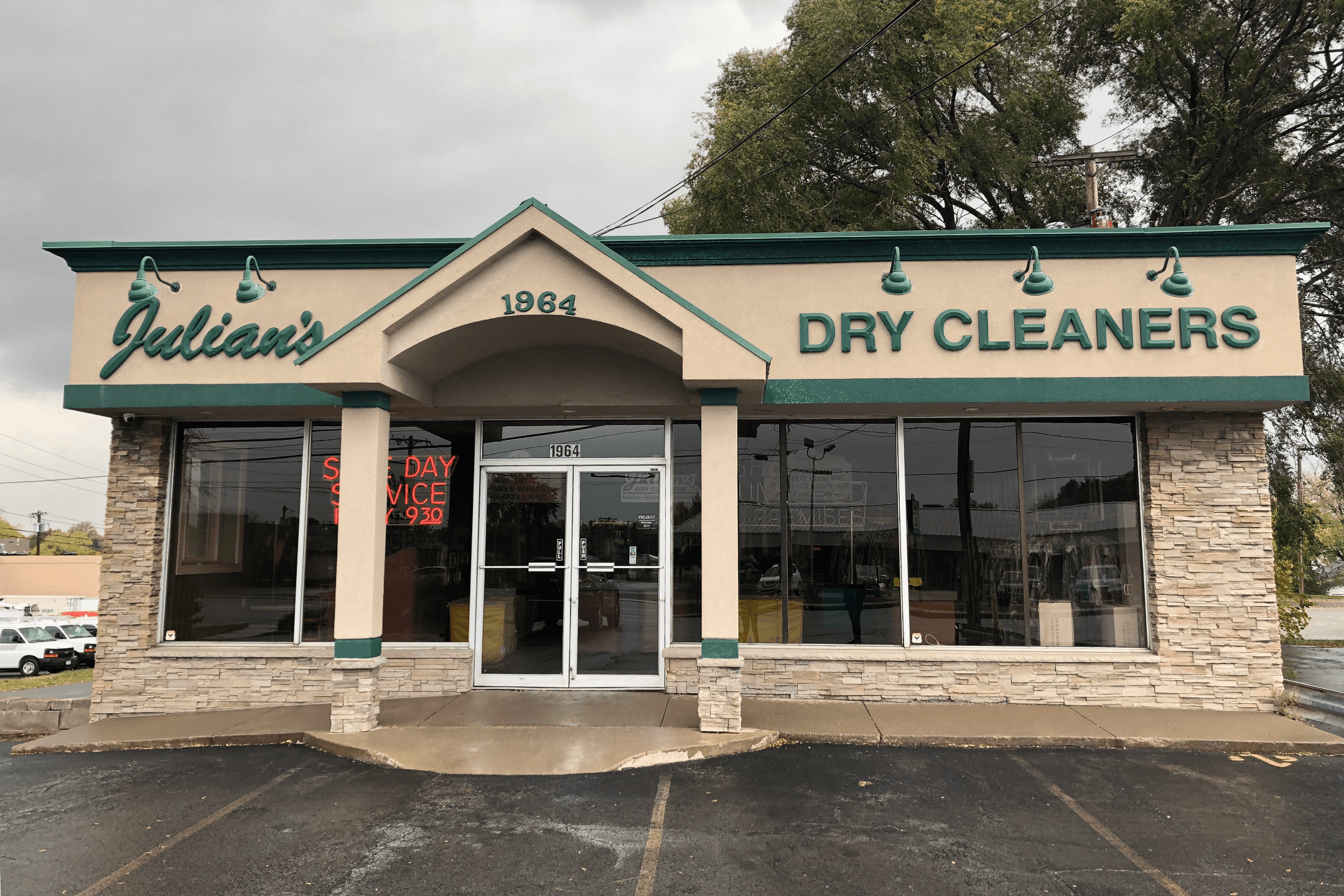 A one-story building with a sign reading "Julian's Dry Cleaners 1964" and a "Same Day Service" neon sign in the window. The exterior features stone accents and large glass windows.