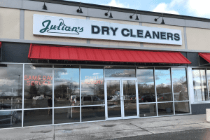 Storefront of Julian&rsquo;s Dry Cleaners with red awnings, glass windows, and neon signs advertising same-day service and shirt laundry.