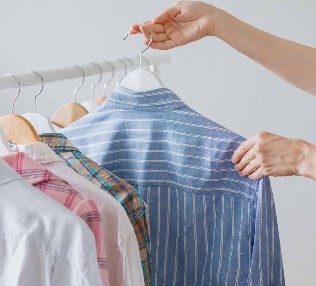 A person selects a blue striped shirt from a clothing rack with several other shirts, all on hangers.