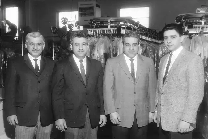 Four men in suits stand side by side inside a dry cleaning shop, with clothes hanging on racks in the background.