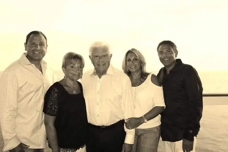 Five adults stand together posing for a photo, with an ocean view in the background. The image is in sepia tone.