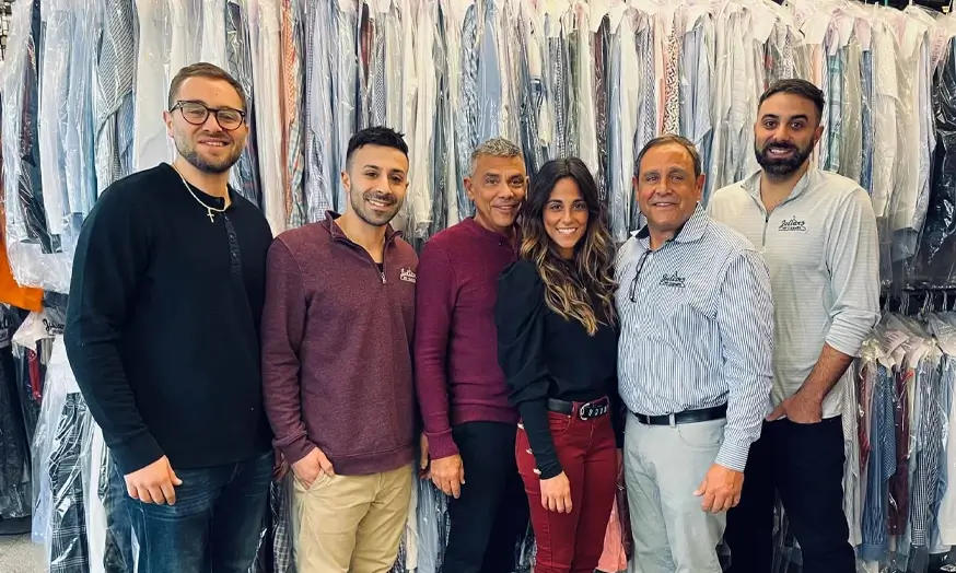 Six people stand in front of a rack of clothes covered in plastic at a dry cleaning business, posing and smiling for the camera.