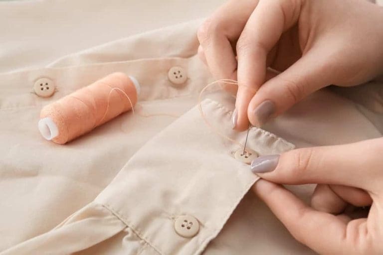 Close-up of hands sewing a button onto a beige shirt with a needle and thread; a spool of matching thread rests on the fabric nearby.