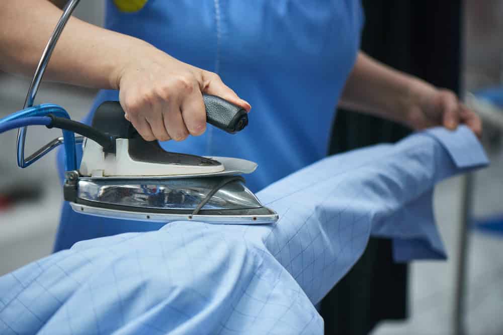 Person ironing a light blue, checkered shirt with a steam iron on an ironing board.