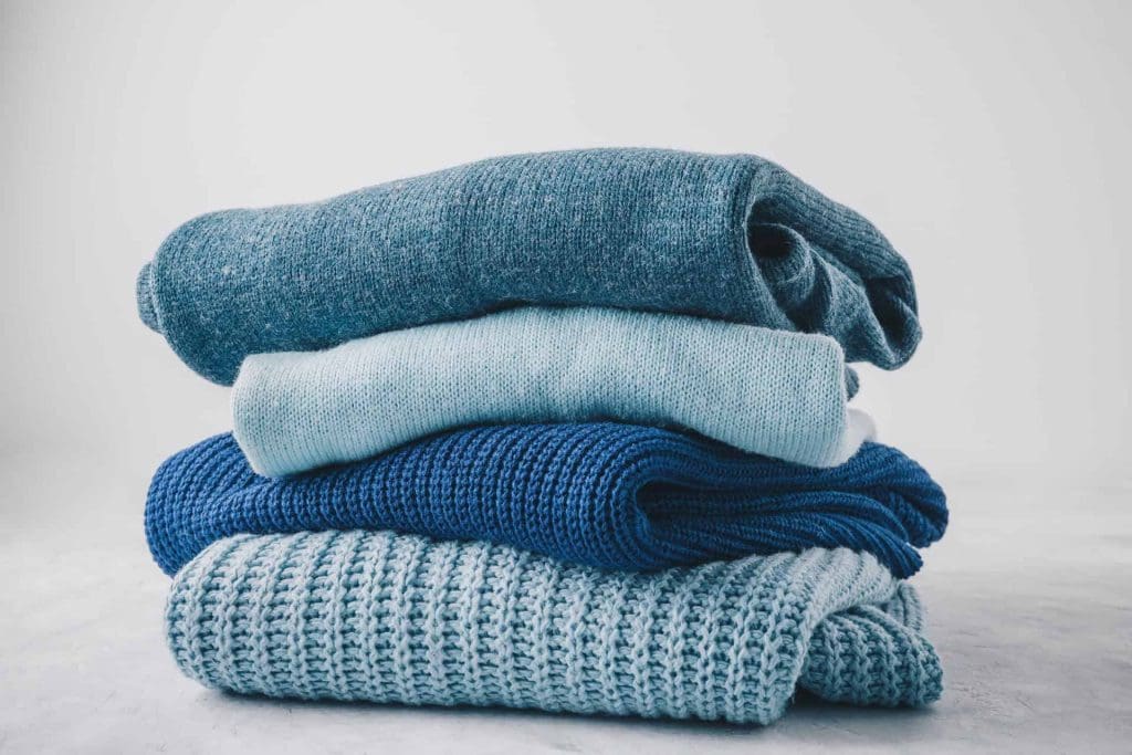 A neatly folded stack of blue and teal sweaters and knitwear is arranged on a light surface against a plain background.