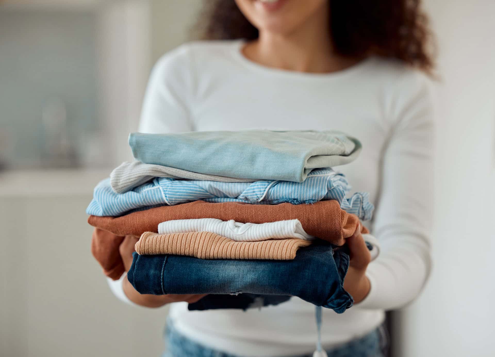 Person holding a neatly folded stack of clothes, including jeans, shirts, and tops, with a blurred background.