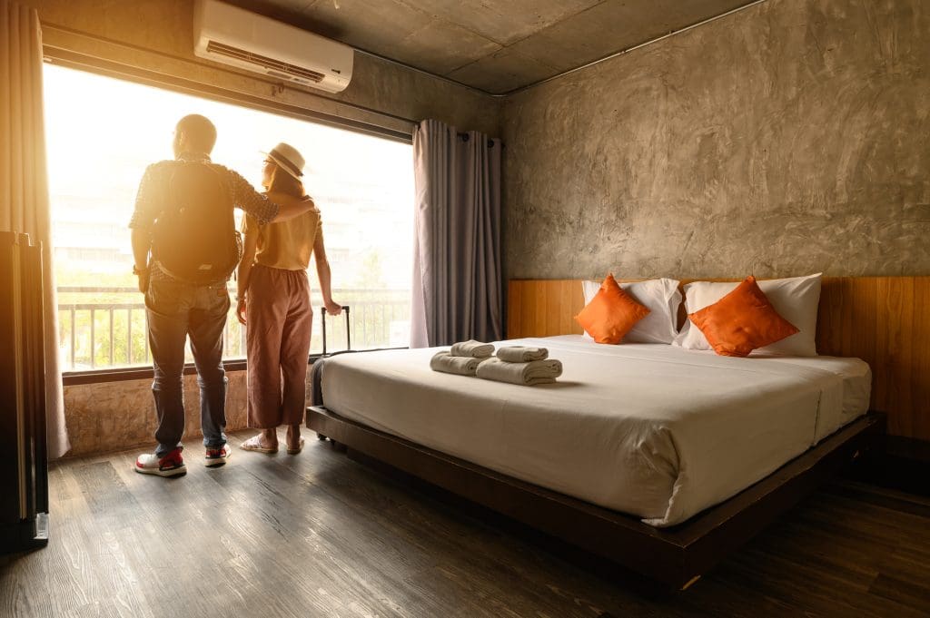 Two people with luggage stand by a large window looking outside in a modern hotel room with a neatly made bed, orange pillows, and folded towels.