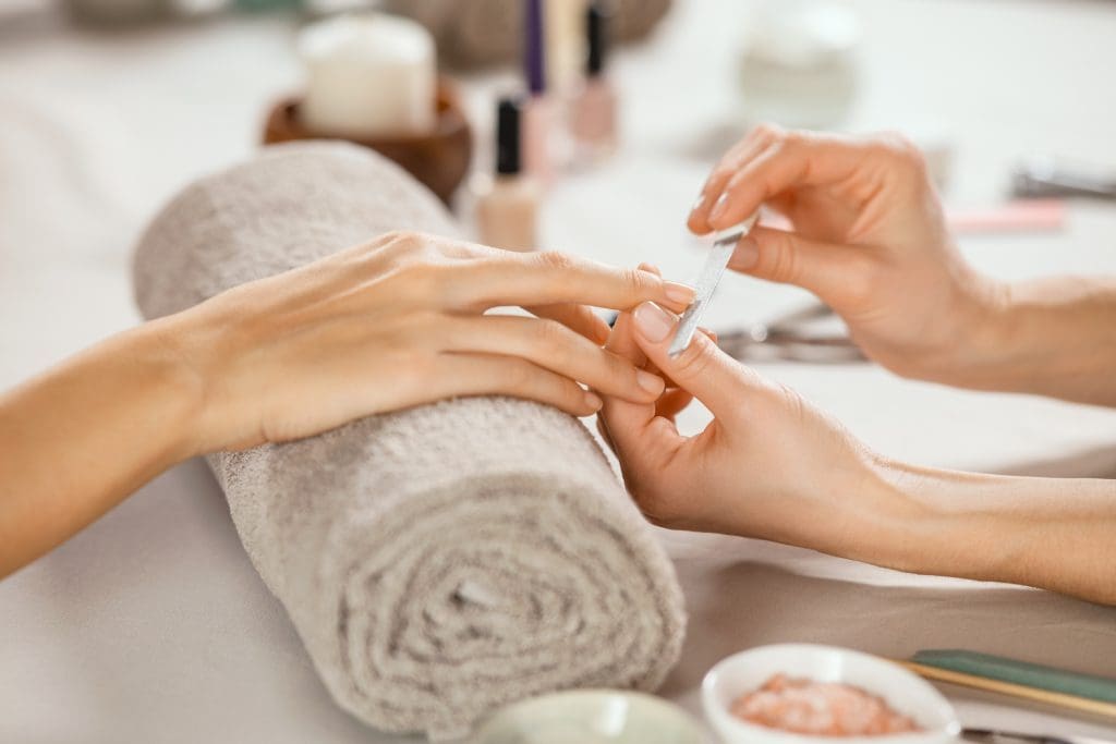 A person receives a manicure as another person files their nails; manicure tools and a rolled towel are visible on the table.