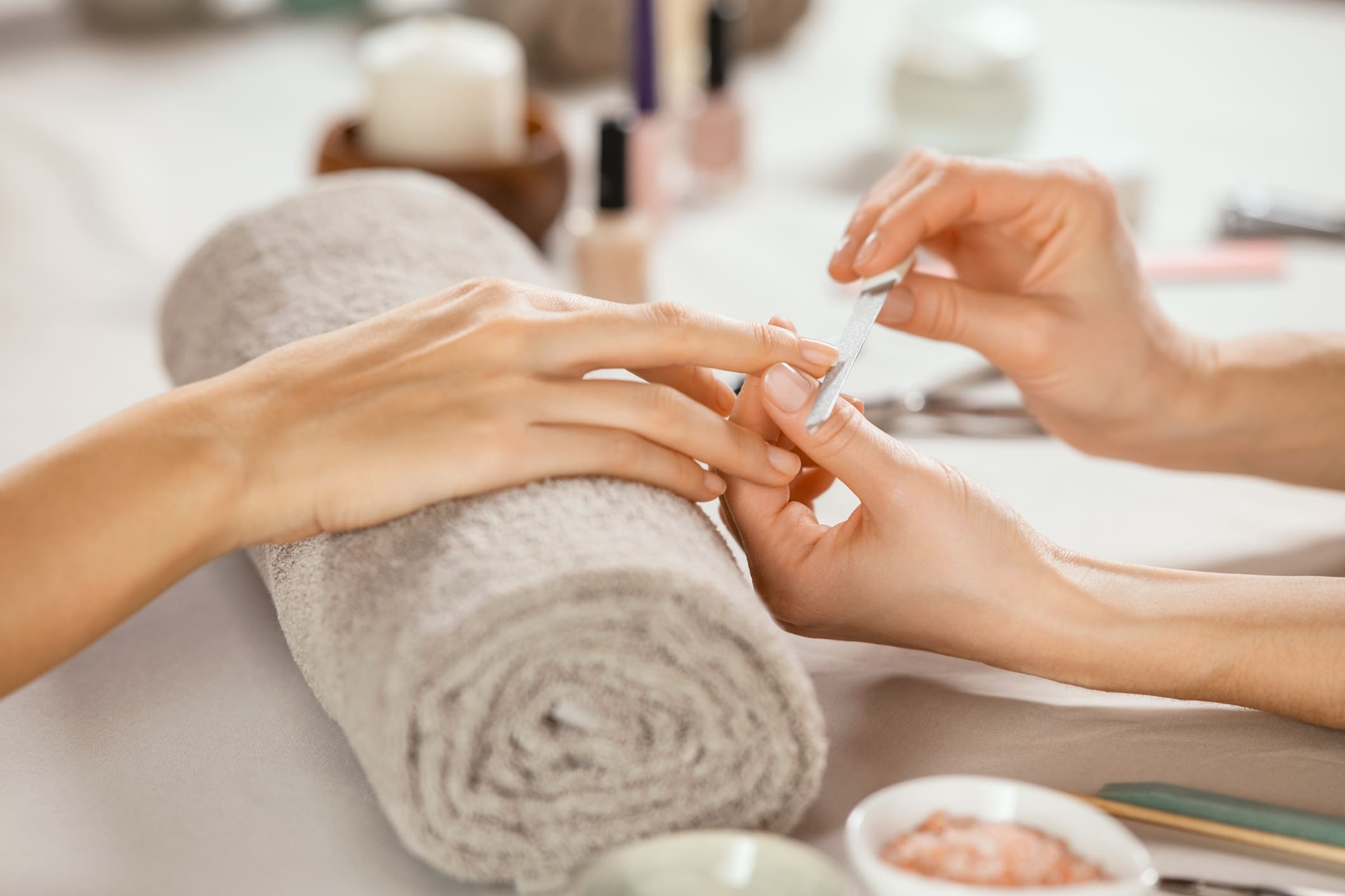 A person receives a manicure as another person files their nails; manicure tools and a rolled towel are visible on the table.