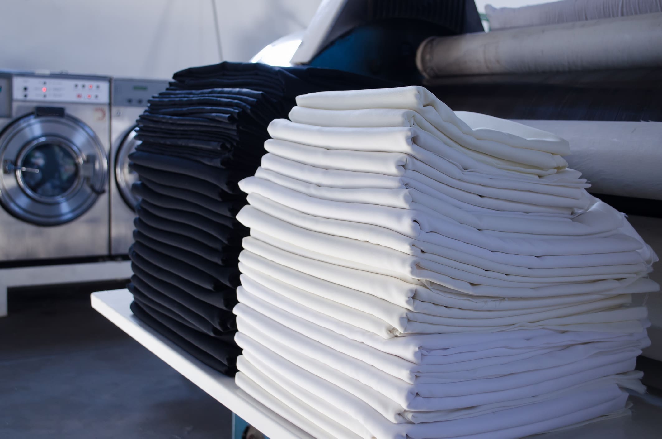 Neatly folded stacks of black and white linens sit on a table in front of industrial washing machines in a laundry facility.