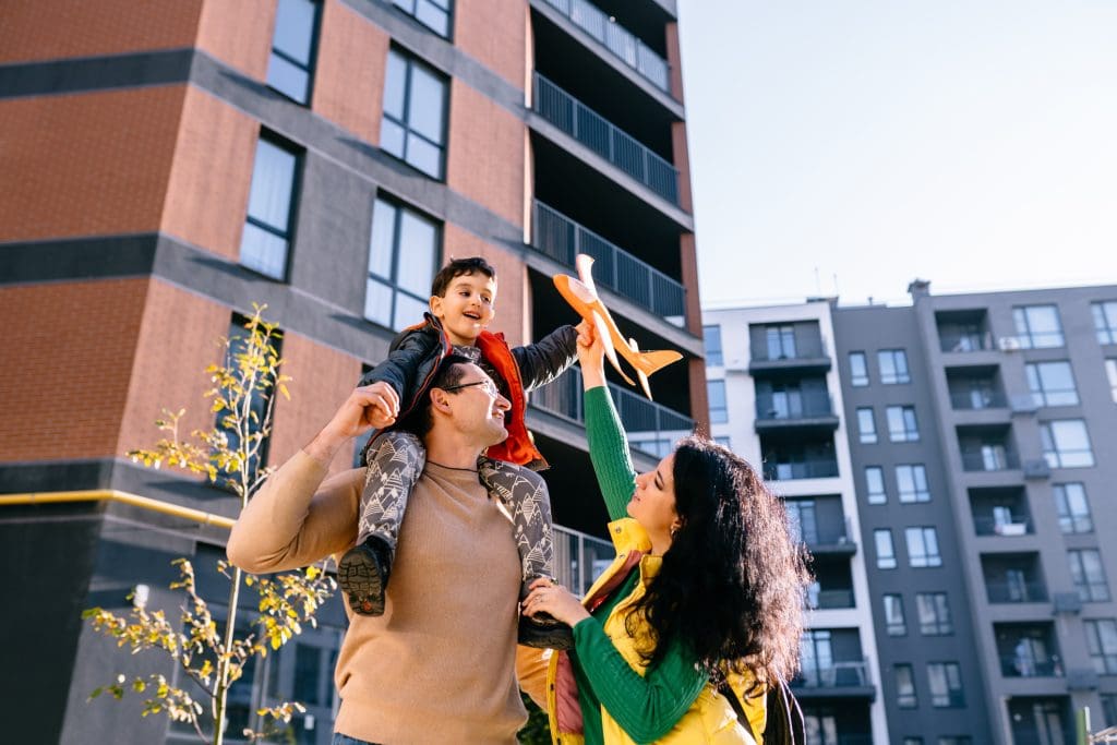 A man carries a smiling child on his shoulders while a woman hands the child an orange toy airplane, with modern apartment buildings in the background.
