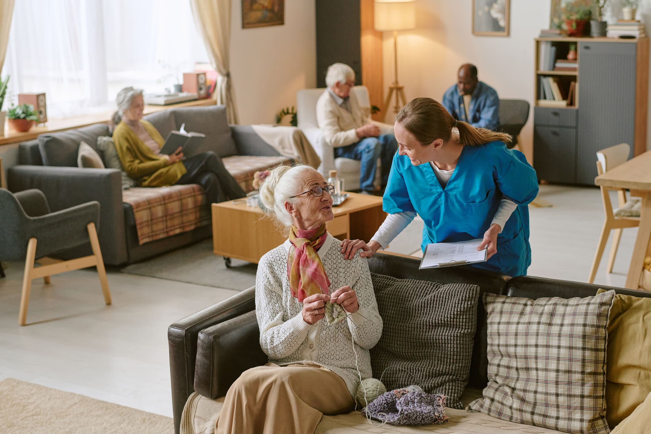 A nurse speaks to an elderly woman knitting on a couch in a common room, while other seniors sit and read in the background.