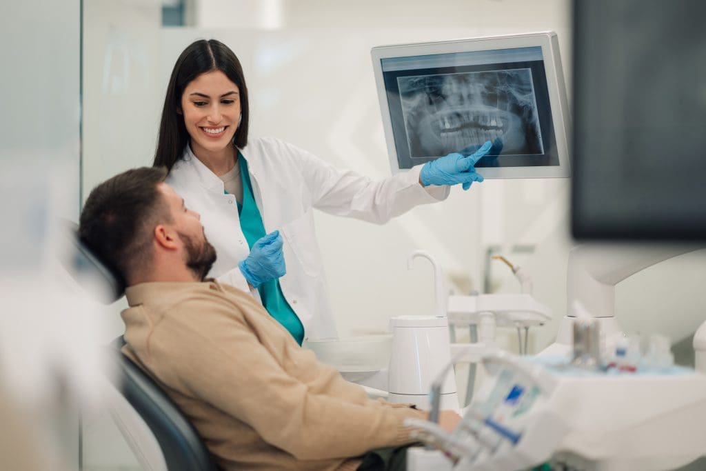 A dentist wearing gloves shows a dental X-ray on a monitor to a patient sitting in a dental chair.