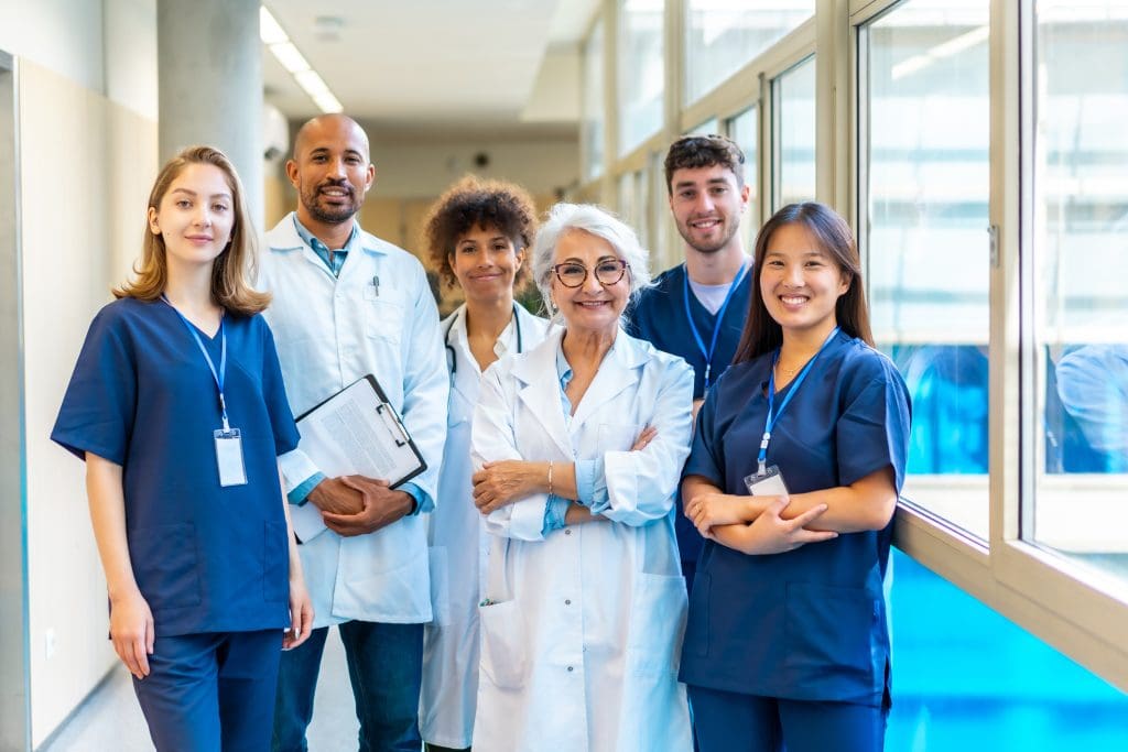 A group of six medical professionals, wearing lab coats and scrubs, stand together in a bright hallway, smiling at the camera.