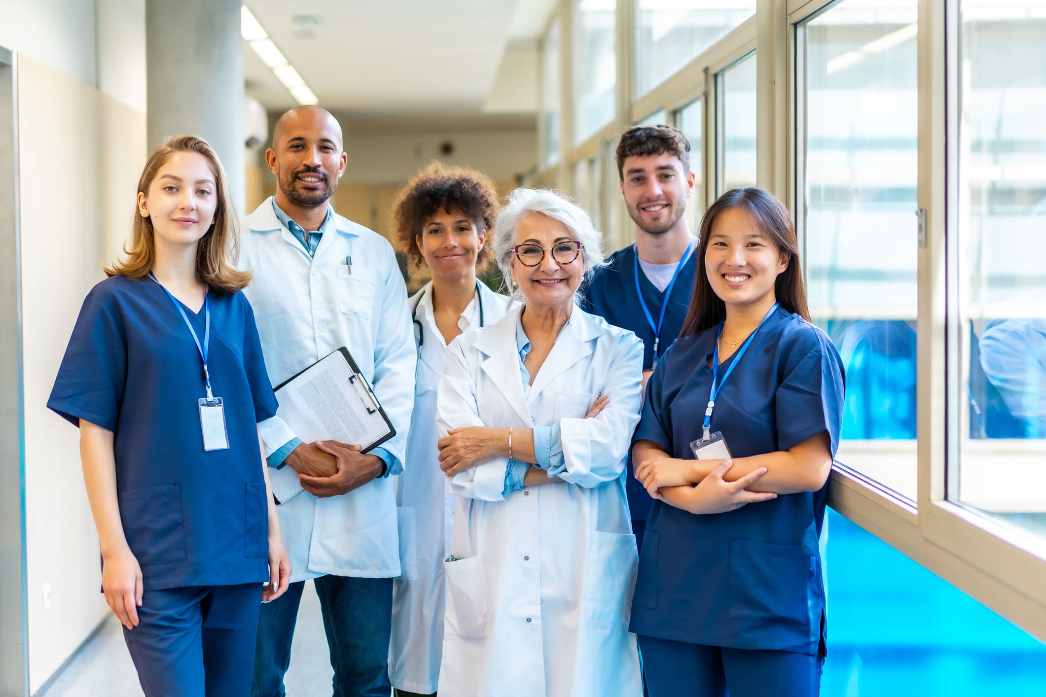 A group of six medical professionals, wearing lab coats and scrubs, stand together in a bright hallway, smiling at the camera.