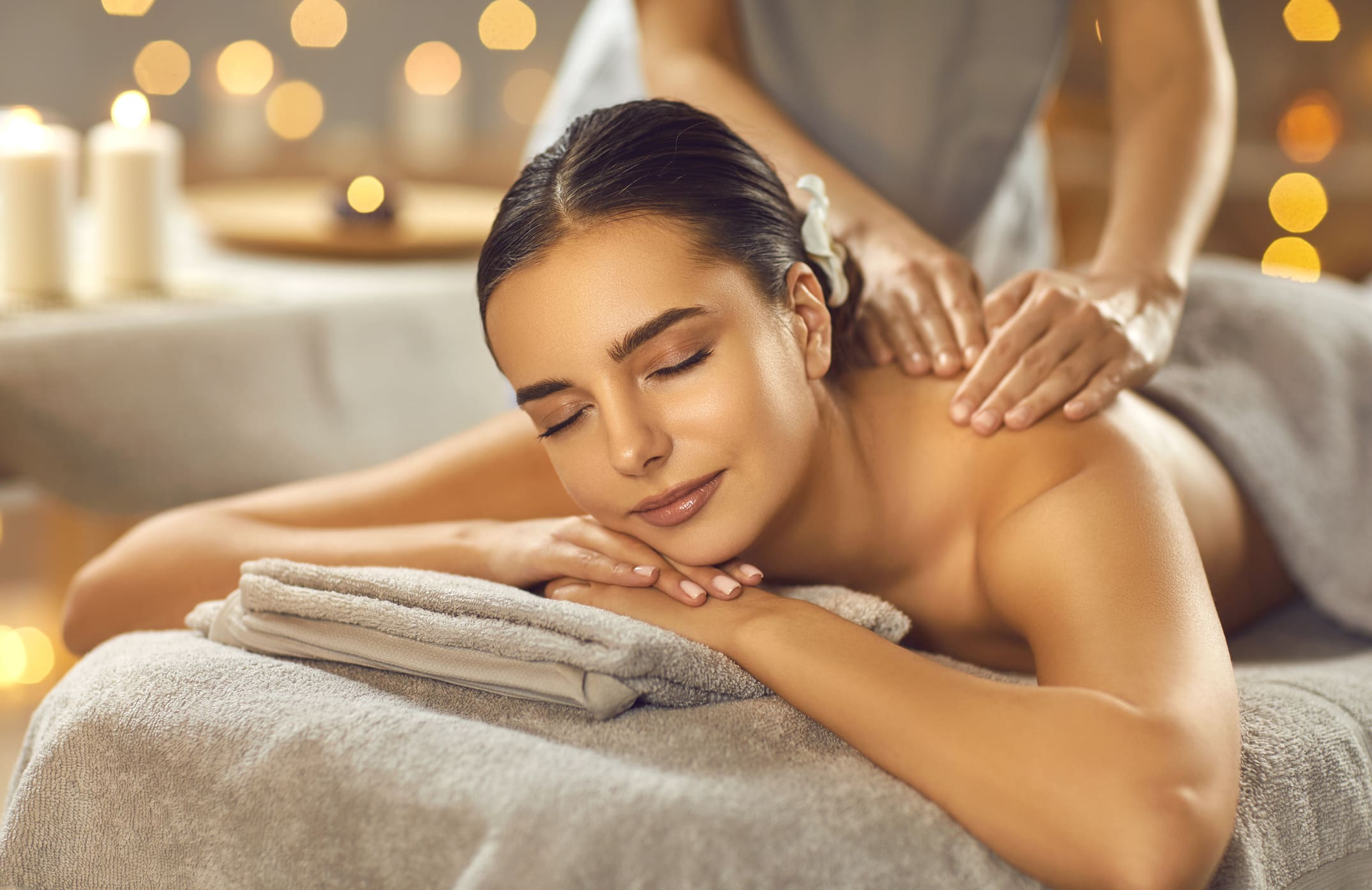 A woman lying on a massage table with her eyes closed receives a back massage in a spa setting with candles and soft lighting in the background.
