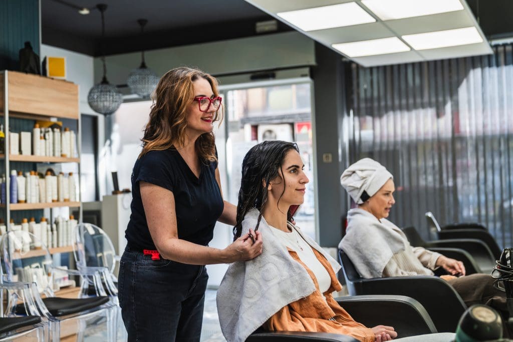 A hairdresser styles a woman's wet hair while another customer with a towel around her head sits nearby in a modern salon.