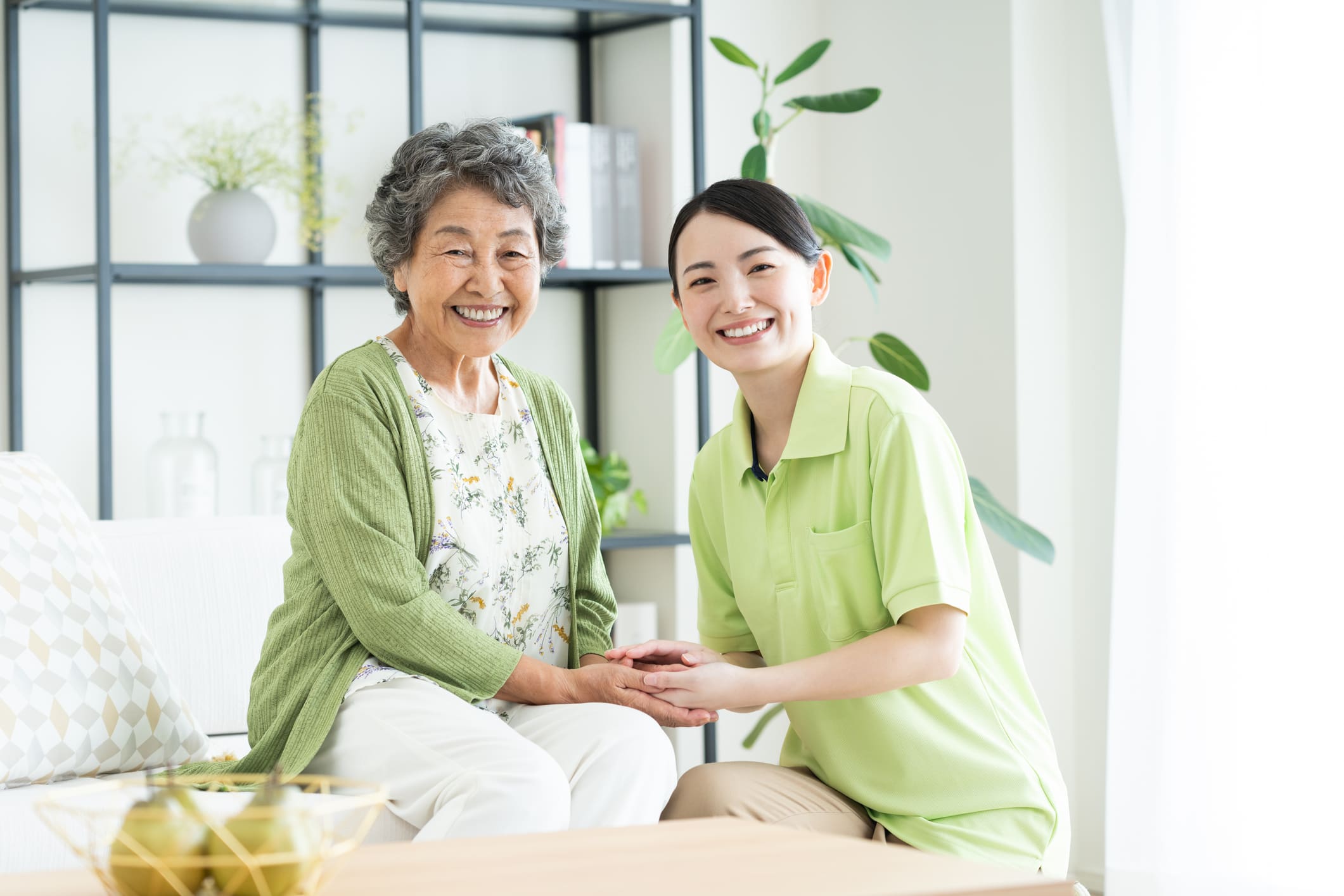 A smiling elderly woman sits on a sofa holding hands with a younger caregiver in a bright, modern living room.