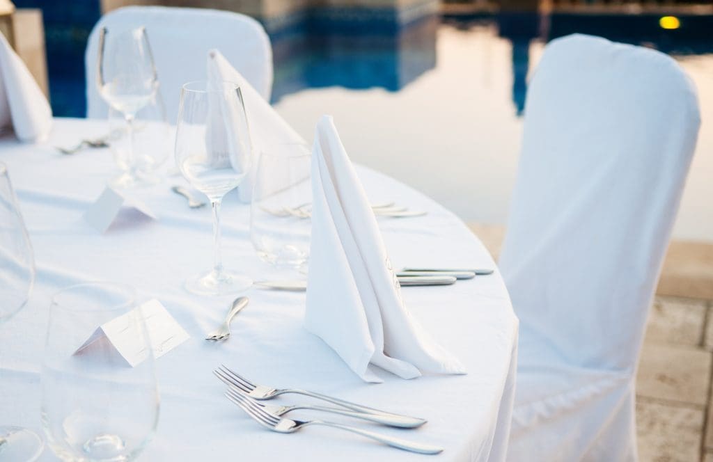 A round table set for a formal event with white tablecloth, folded white napkins, glassware, cutlery, and name cards by a poolside.