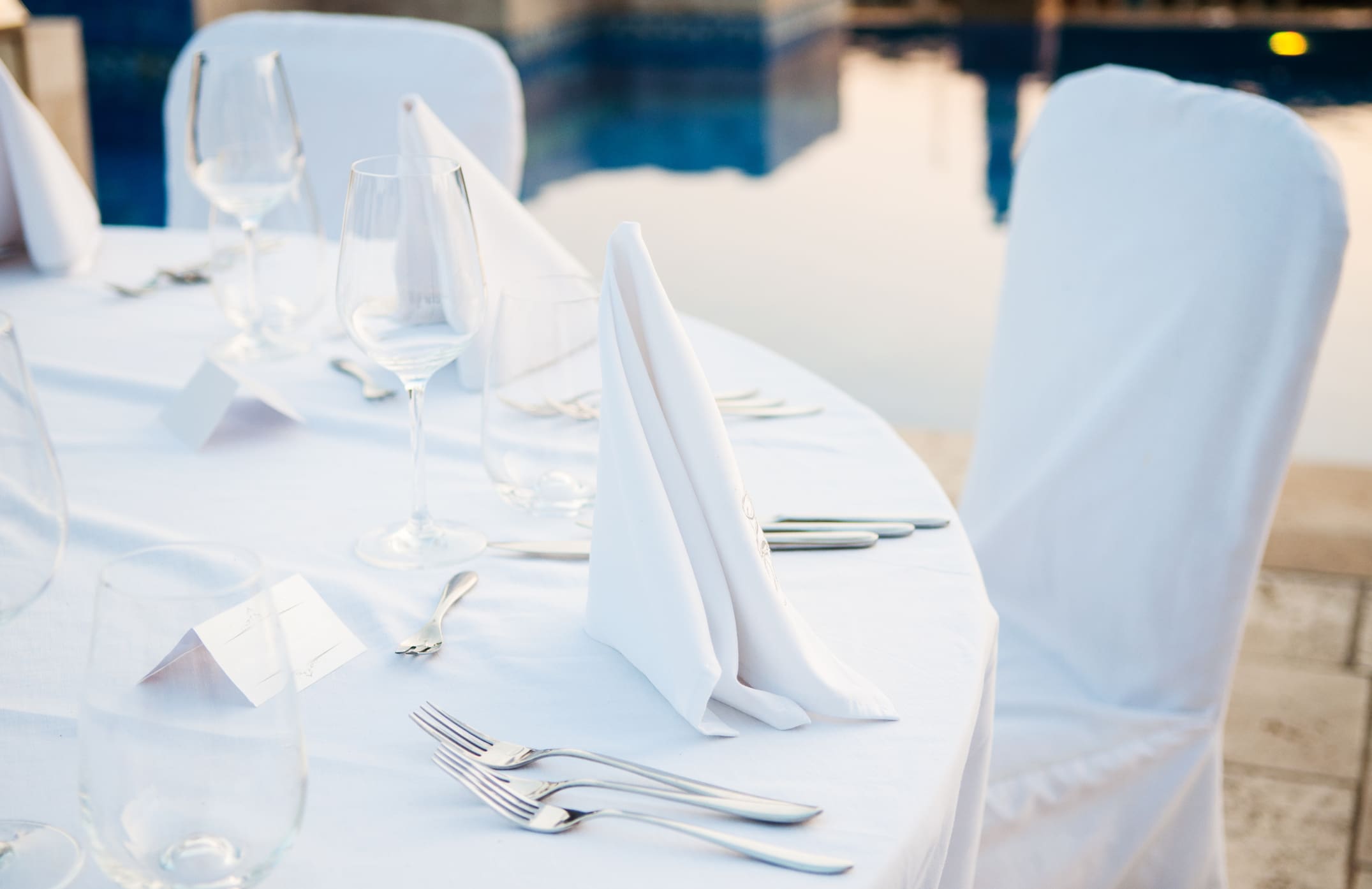 A round table set for a formal event with white tablecloth, folded white napkins, glassware, cutlery, and name cards by a poolside.