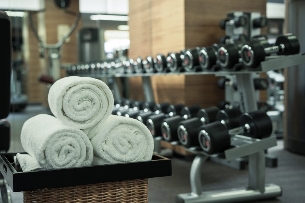 A basket of rolled white towels sits in front of racks filled with black dumbbells in a gym setting.
