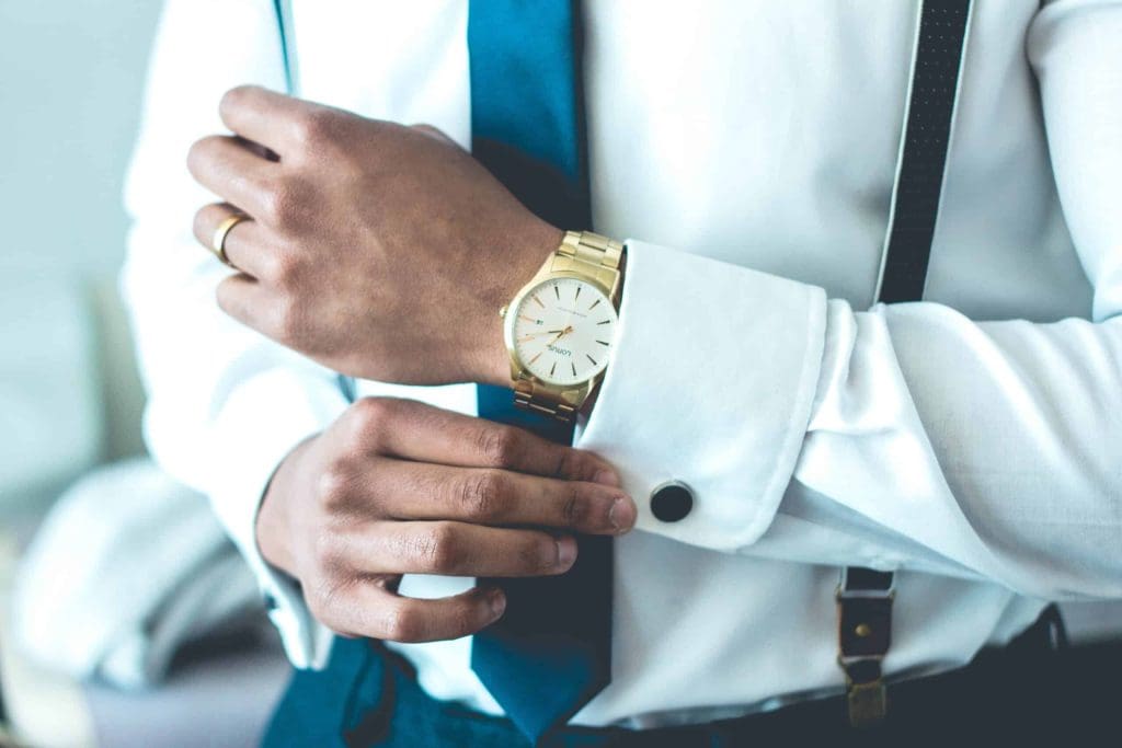 A person wearing a white dress shirt, blue tie, and suspenders adjusts their gold wristwatch.