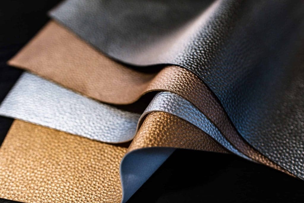 Several sheets of textured metallic leather in shades of gold, silver, and bronze are layered and slightly overlapping on a dark surface.