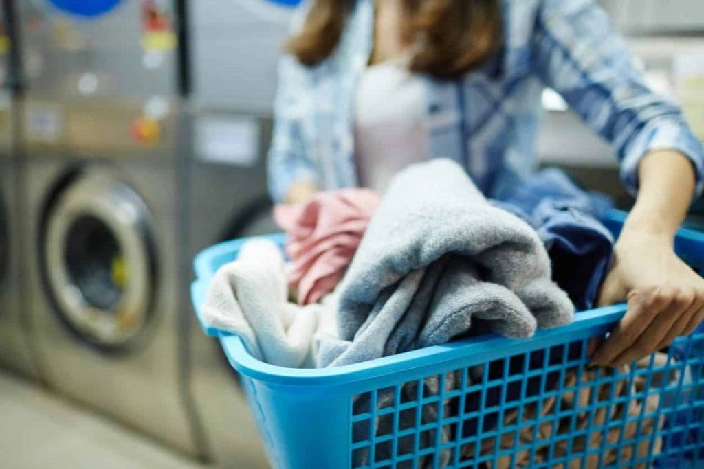 Person holding a blue laundry basket filled with clothes in front of washing machines at a laundromat.