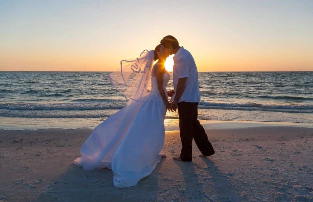 A bride and groom stand on a beach at sunset, holding hands and touching foreheads, with the ocean and sun in the background.